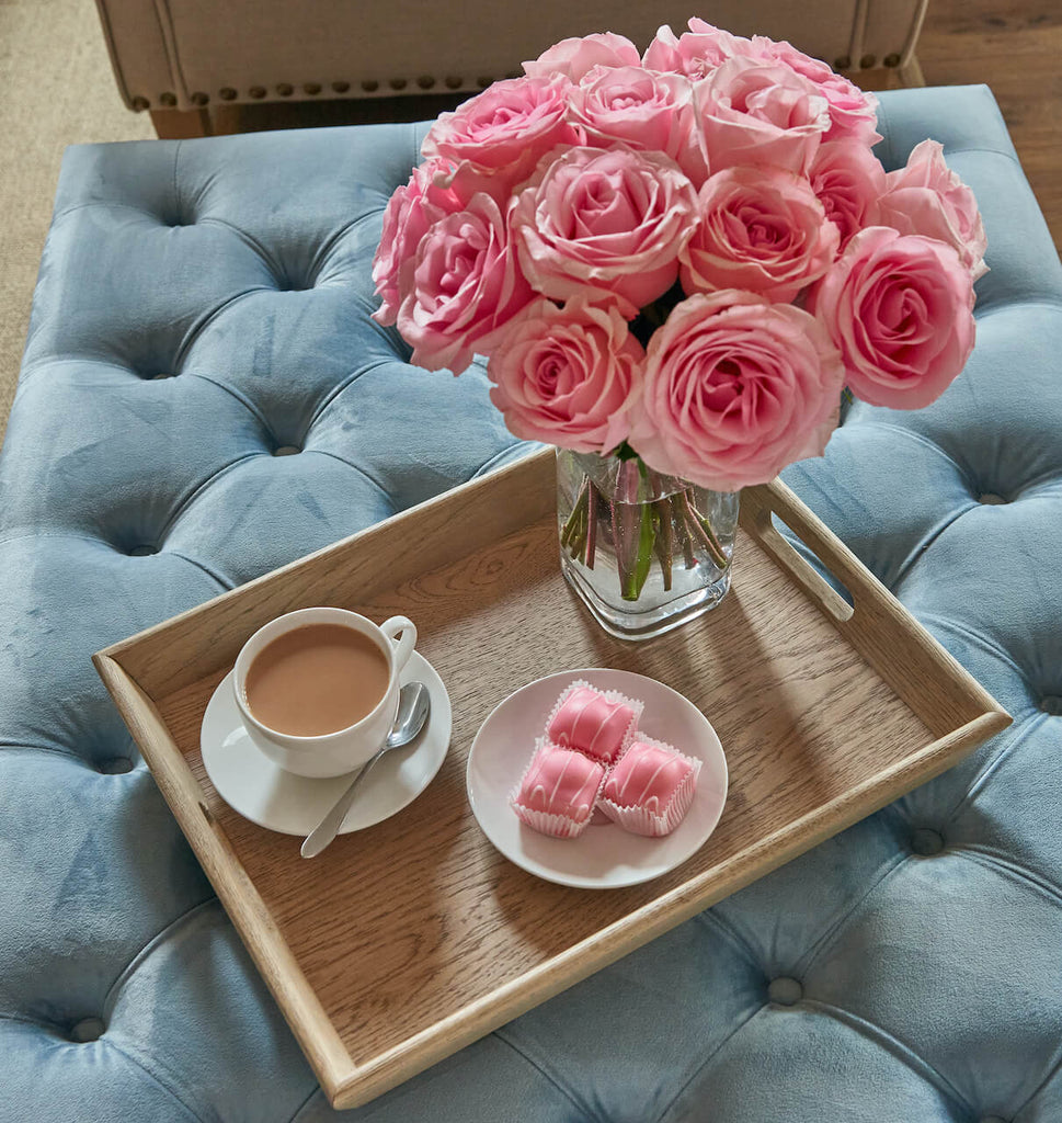 Weathered oak tray on pale blue ottoman with pink roses, pink fondant cakes and cup of tea on saucer